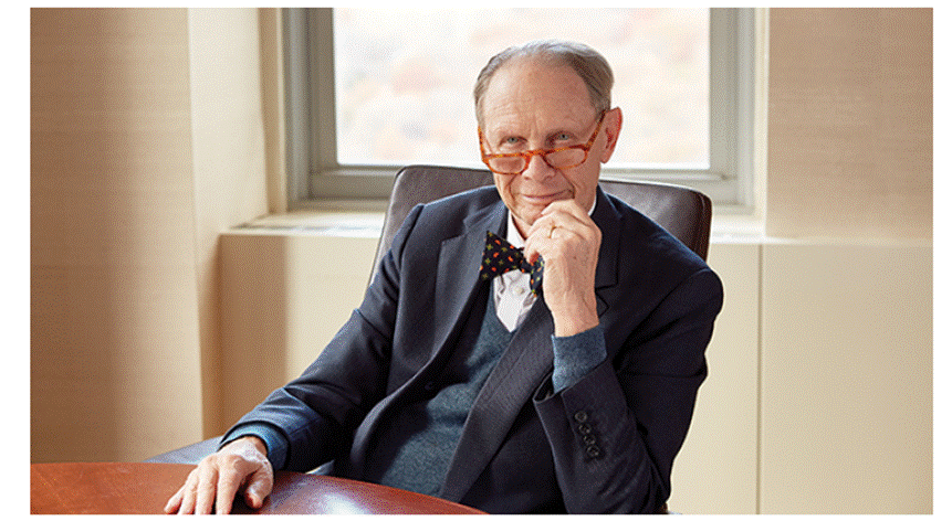 A person in a suit and bow tie sitting at a table

Description automatically generated